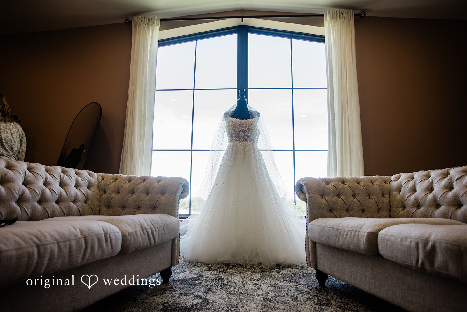 A wedding dress hanging elegantly in front of a bright window framed by two indoor couches.