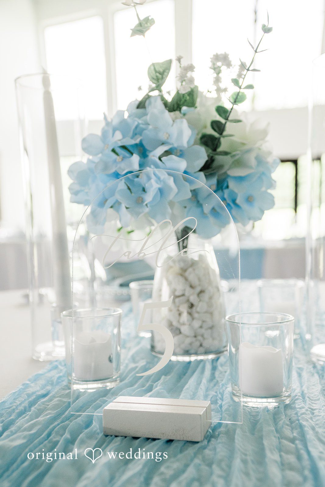 A wedding reception table setting featuring a light blue floral centerpiece and a white table runner.