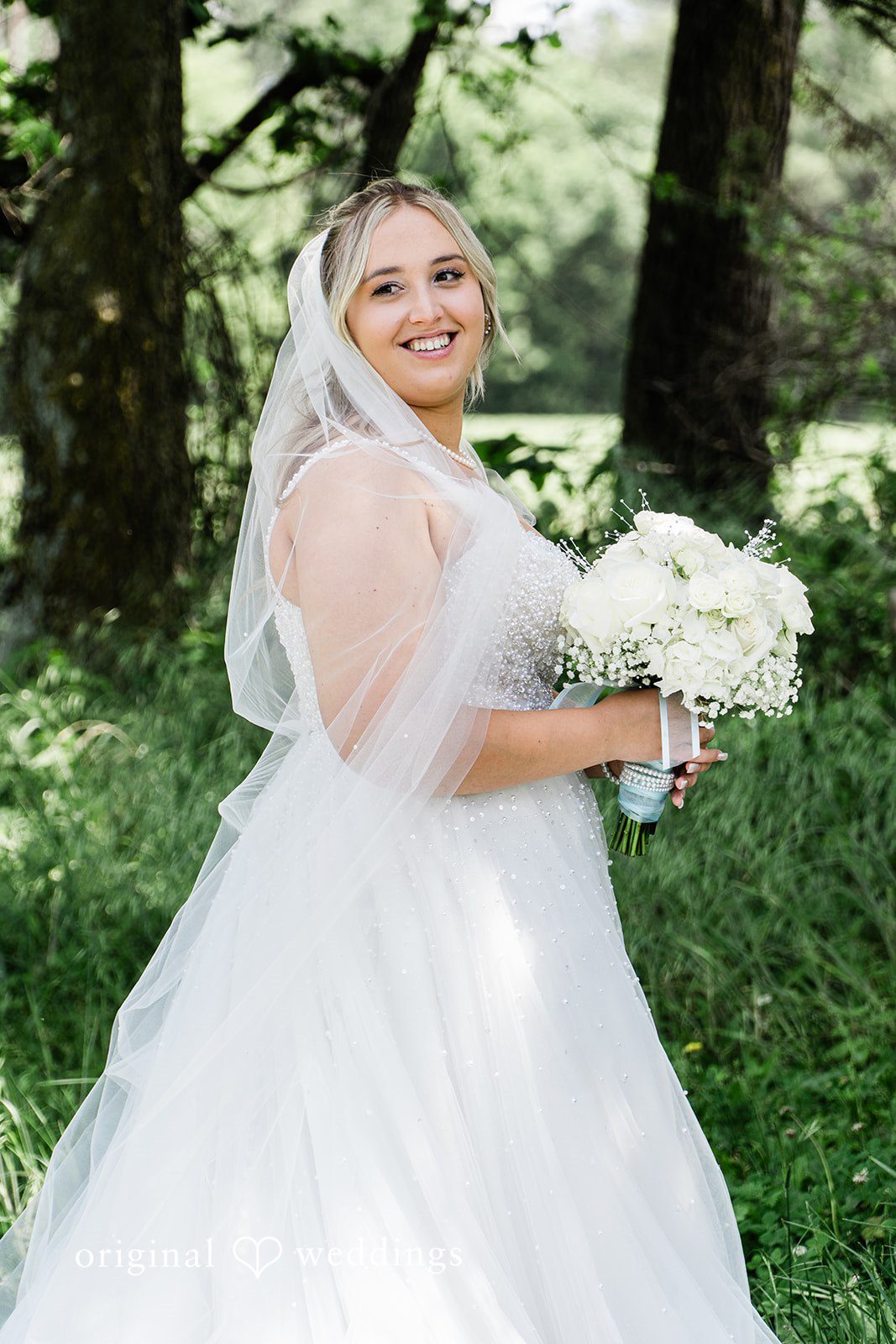 A bride smiling while holding a white bouquet in front of dense green trees.