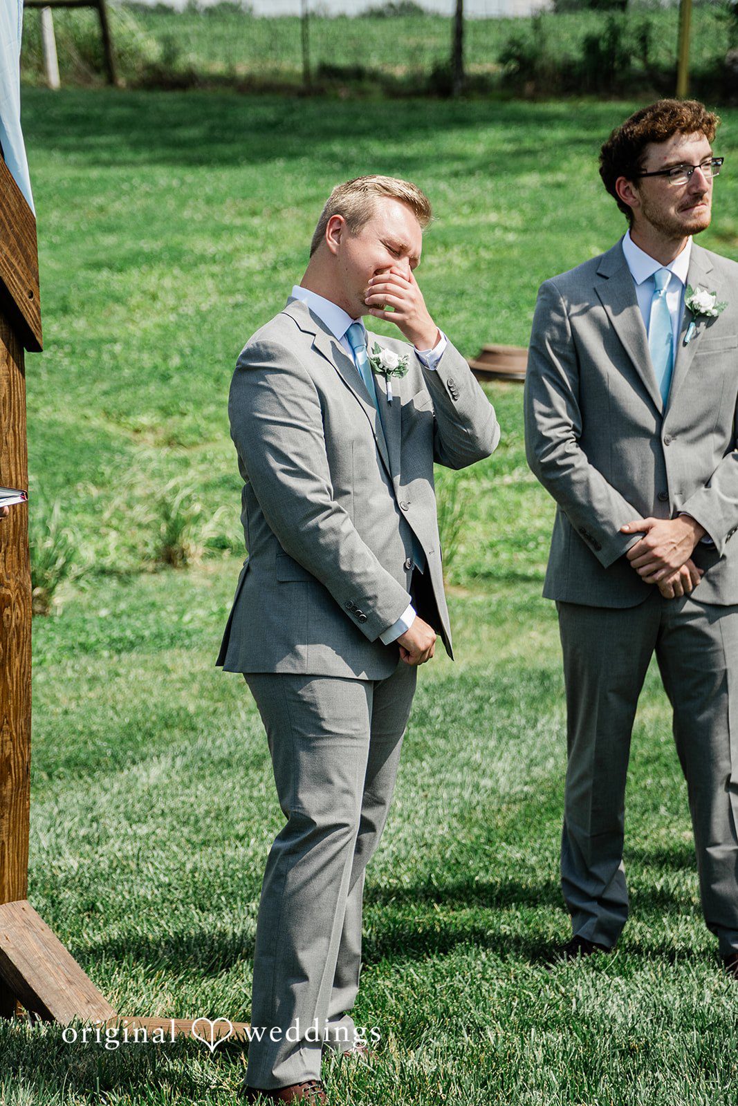 A groom and a groomsman standing together in matching grey suits on an open lawn.