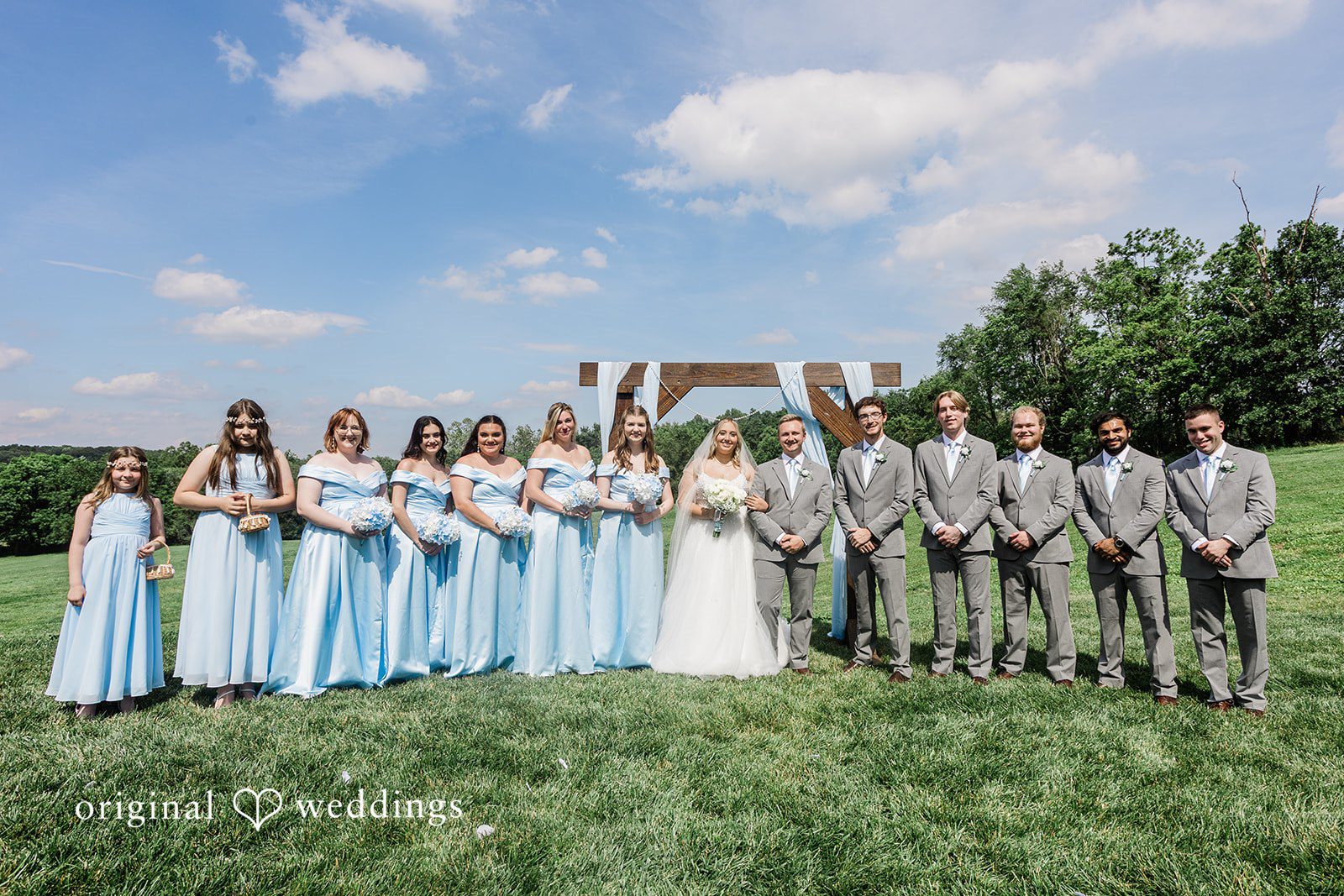 A large wedding party lined up in a grassy field for a group photograph.