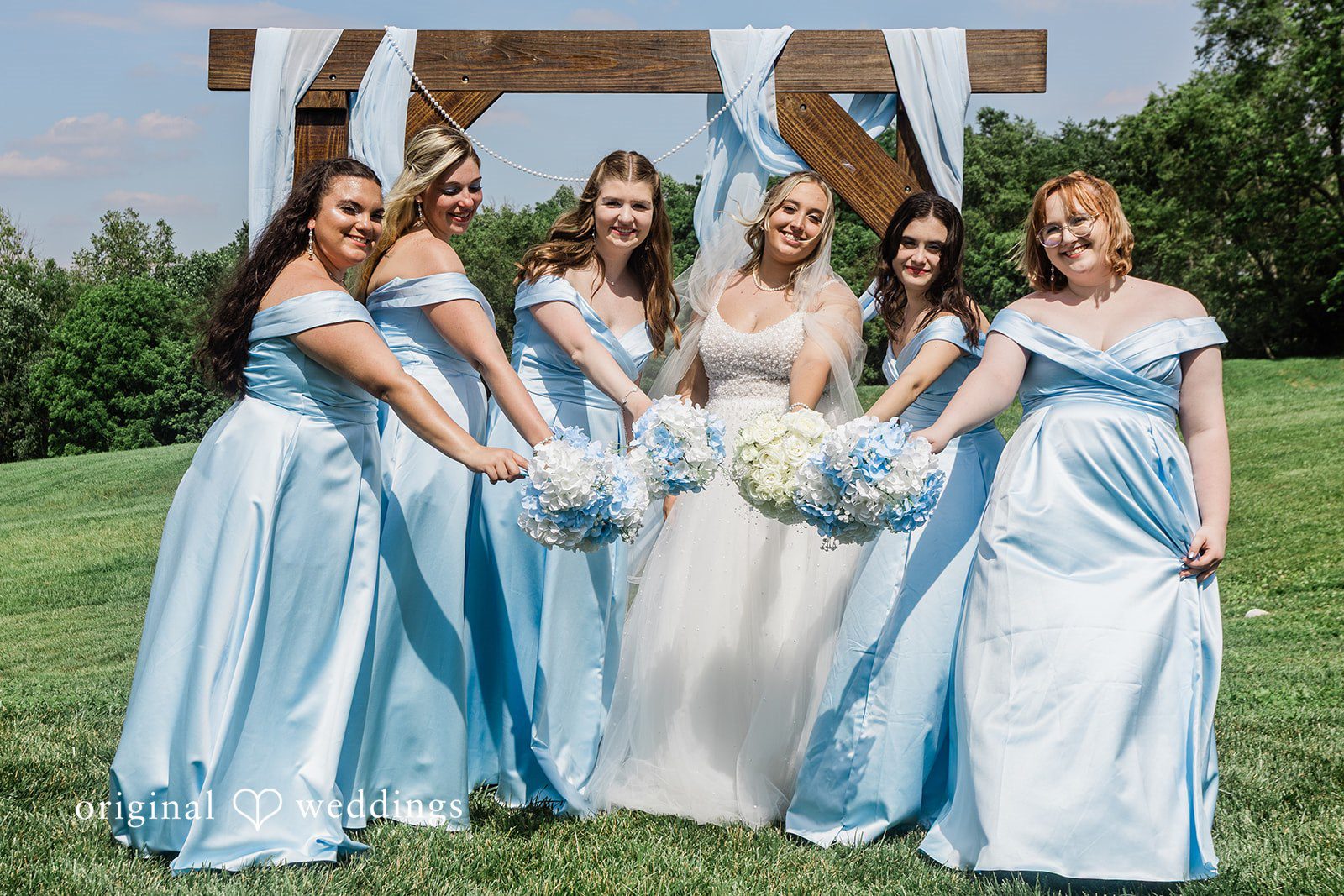 Bride and bridesmaids in light blue dresses posing together under a rustic wooden structure.