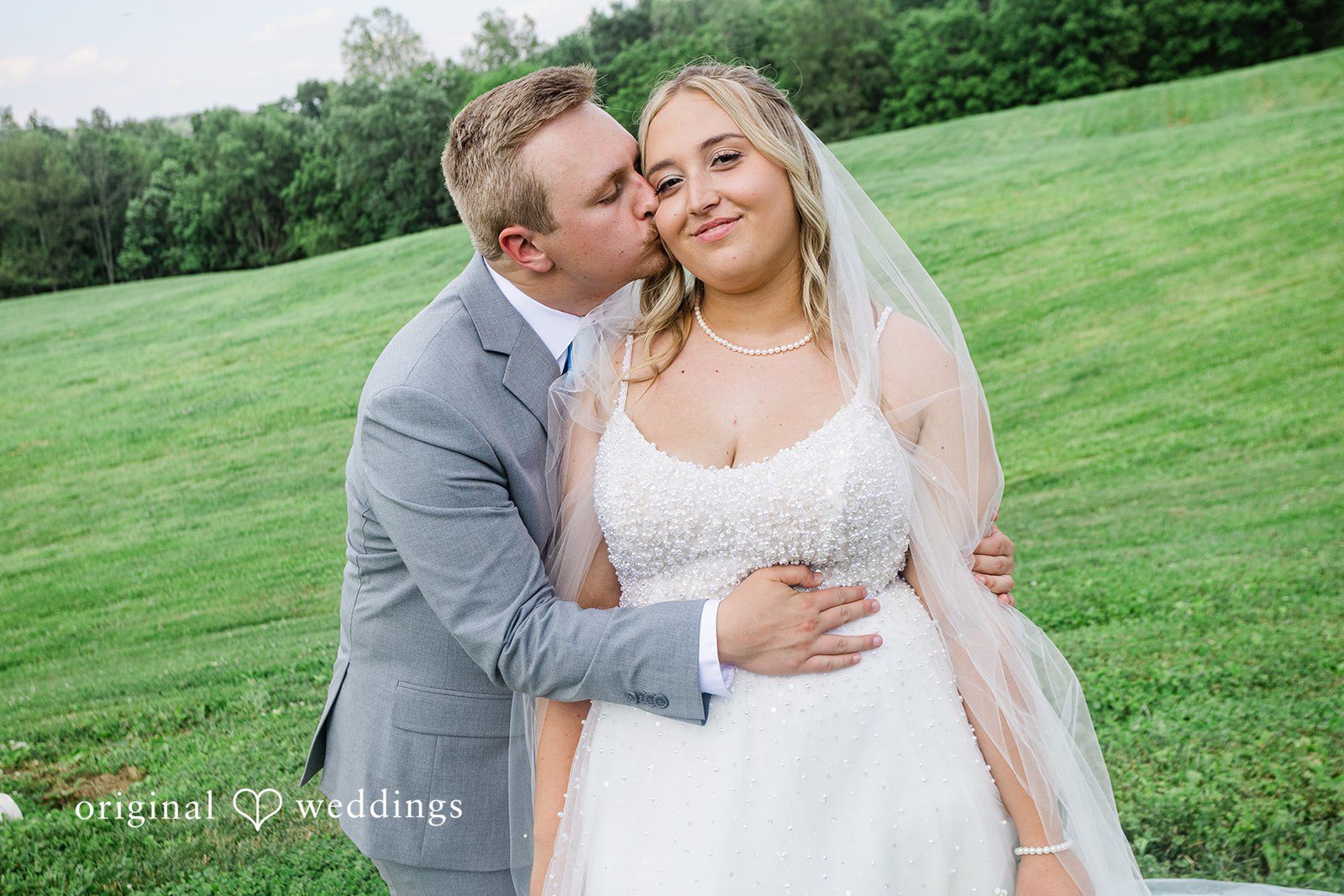A bride and groom embracing on a grassy slope while smiling.