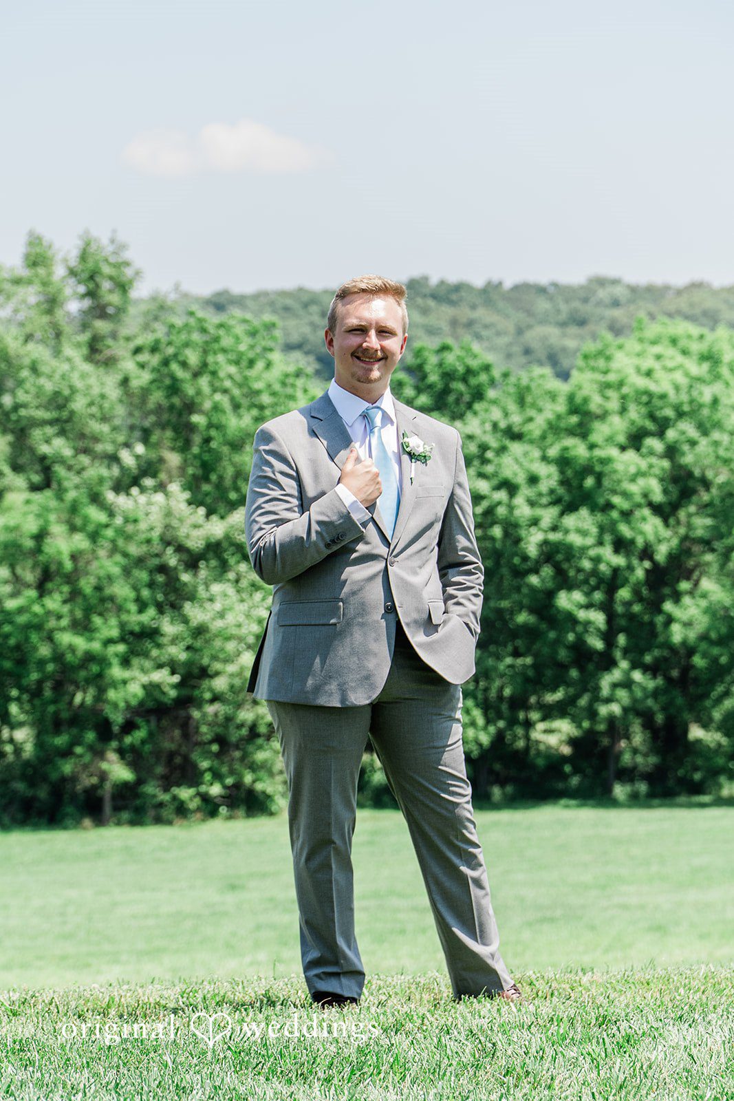 A groom standing alone on a grassy hill with a background of lush green trees.