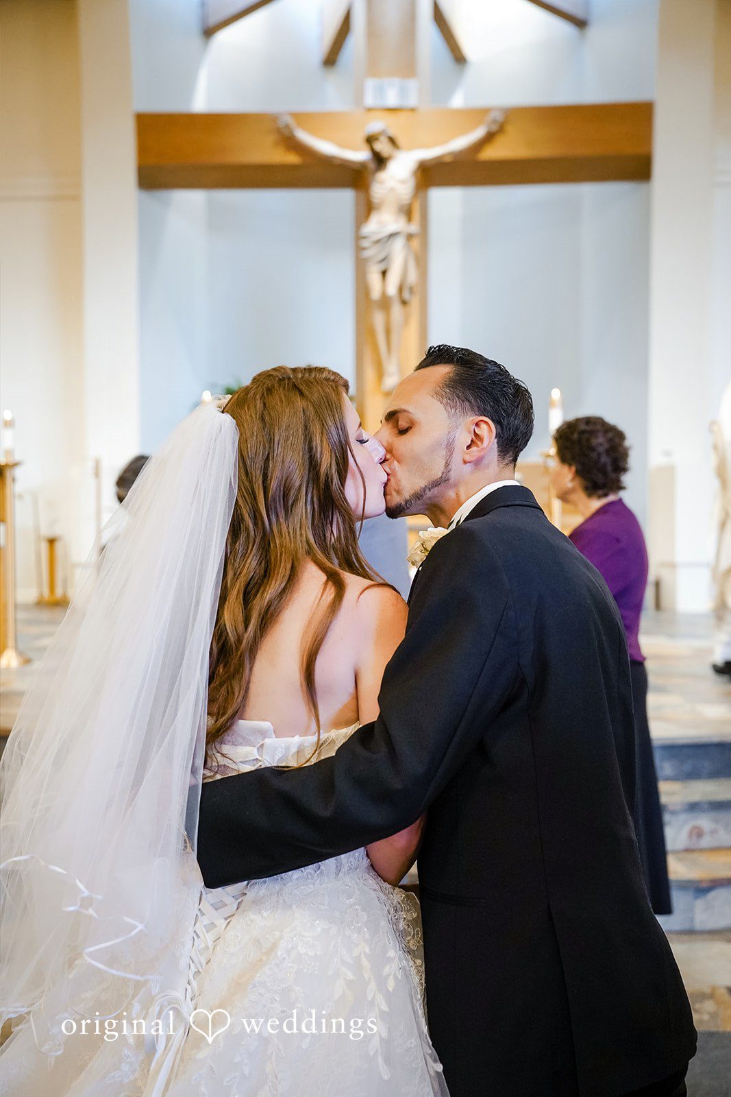 Vincenzo + Kristin A bride with a long veil and a groom in a black suit share a kiss during their wedding ceremony in front of a church altar with a crucifix.