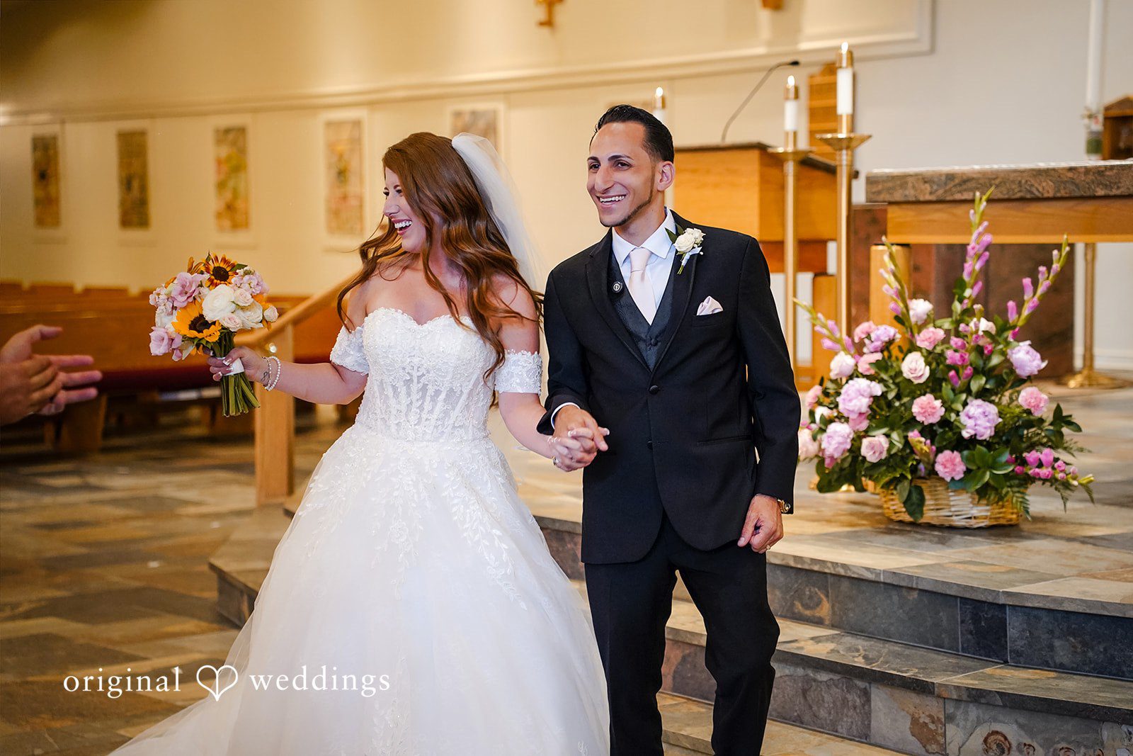 Vincenzo + Kristin A smiling bride and groom hold hands as they walk down the aisle together after their church wedding ceremony.