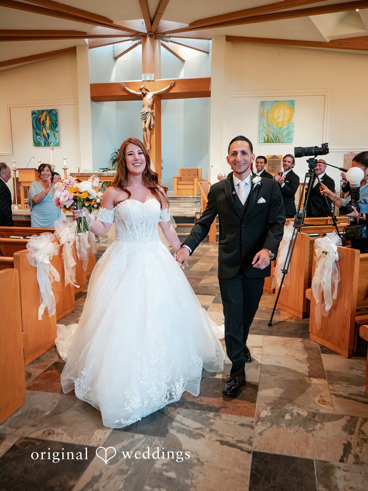 July 11, 2026 – Vincenzo & Kristin – (ID-4669) REVISED 9.6 A smiling bride and groom hold hands as they walk down the aisle inside a church following their wedding ceremony.