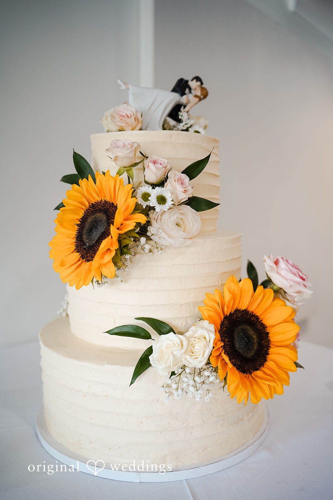 Vincenzo + Kristin A multi-tiered white wedding cake decorated with fresh sunflowers.