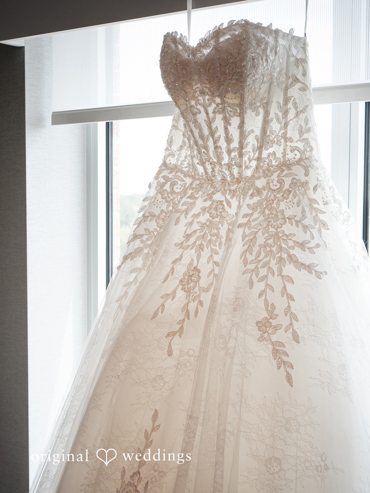 Vincenzo + Kristin An elegant white wedding dress hanging in front of a bright window.