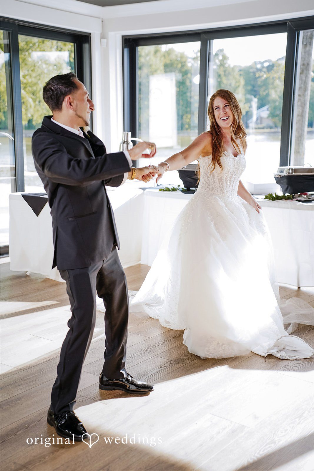 Vincenzo + Kristin A laughing bride and groom hold hands while dancing together.