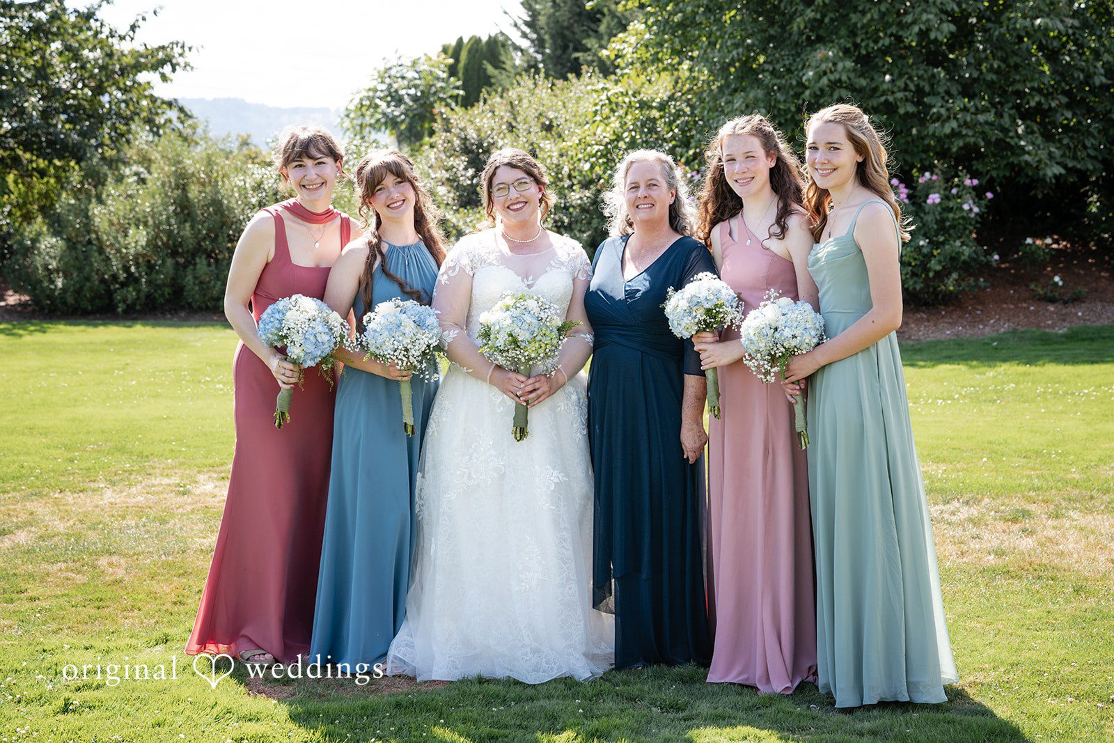 Emma + Aidan Bride and bridesmaids wearing mismatched blue and pink dresses standing together on a lawn.