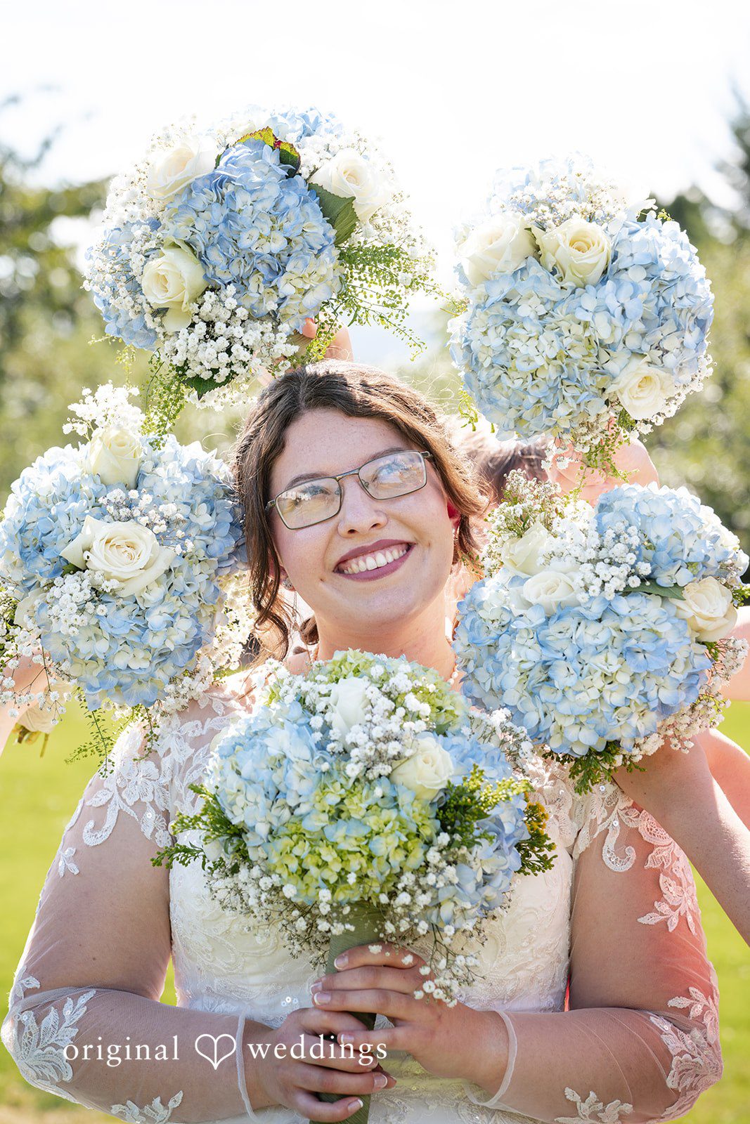 Emma + Aidan A close-up of a smiling bride holding a large bouquet of light blue hydrangeas.