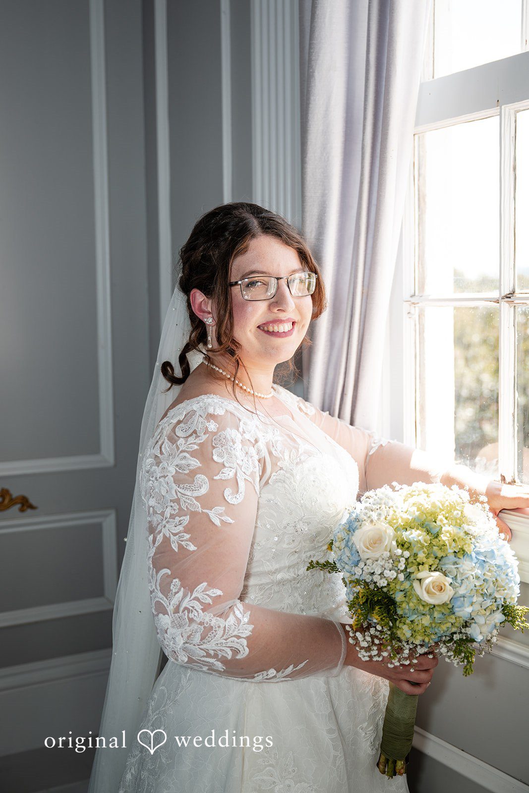 Emma + Aidan A smiling bride looking out a window while holding a bouquet of white flowers.