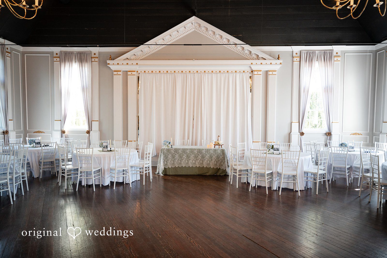 Emma + Aidan An empty, elegant white reception hall filled with round tables and chairs.