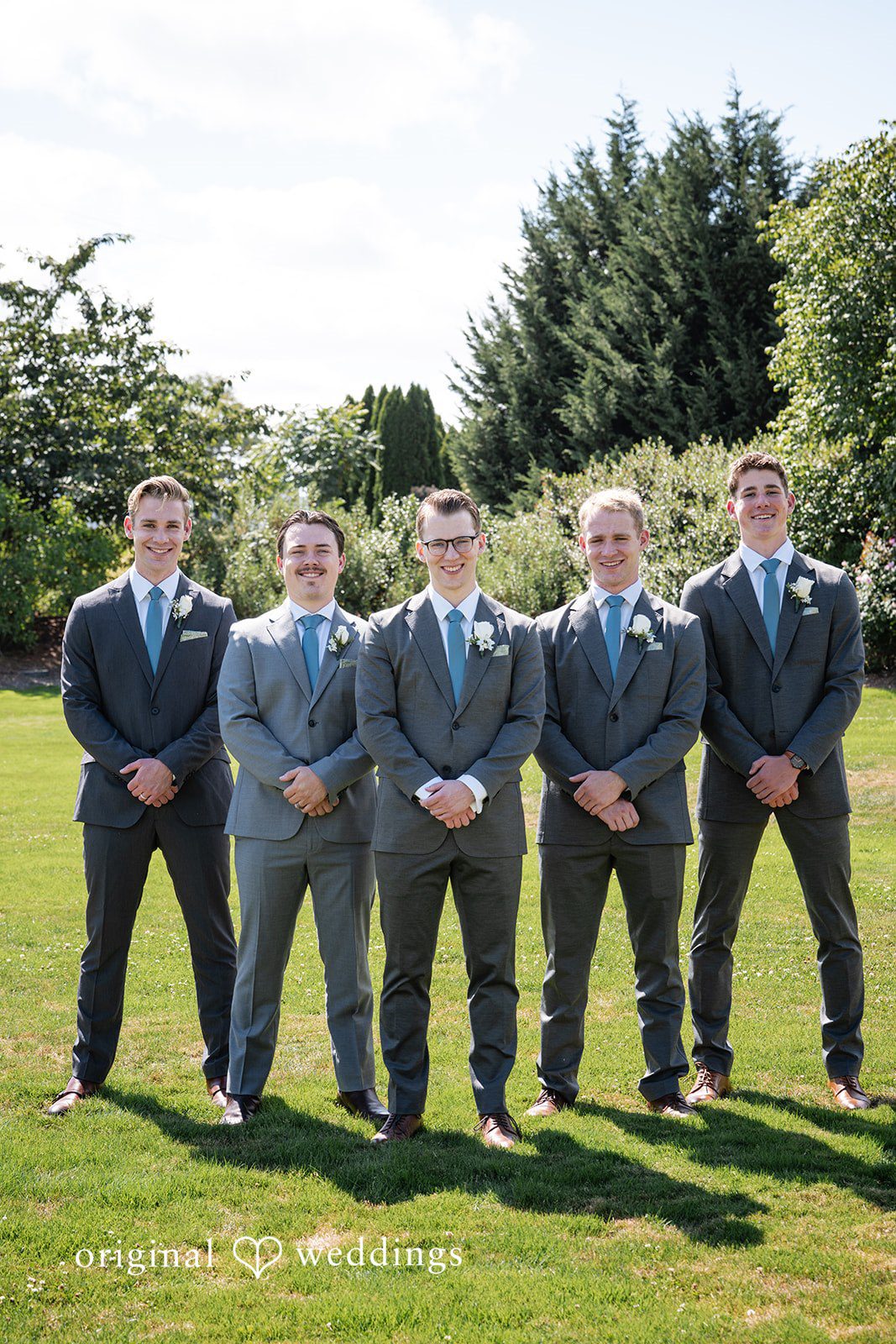 Emma + Aidan Groom and groomsmen in matching grey suits posing together on a grassy field.
