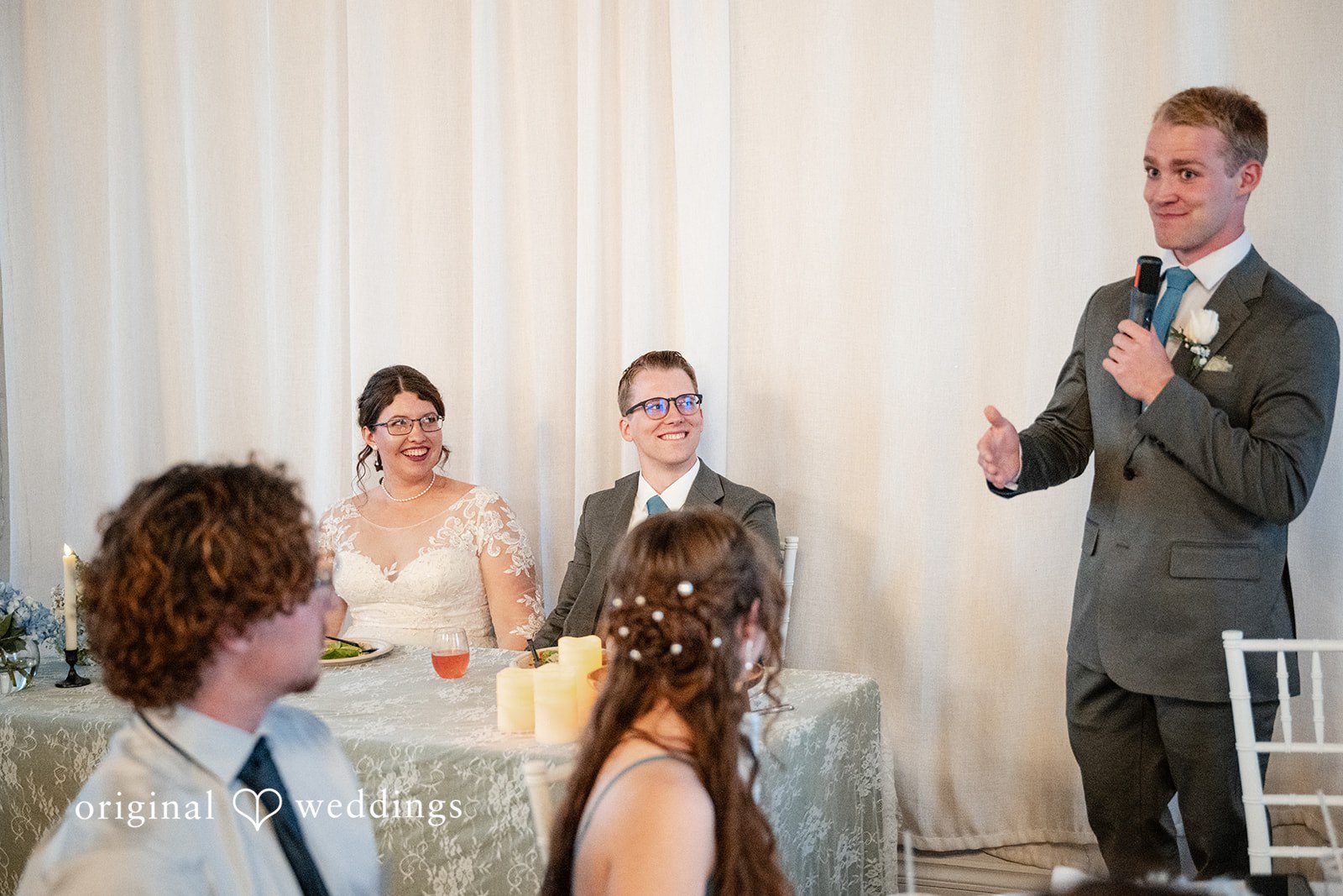 Emma + Aidan A man in a suit speaking into a microphone while wedding guests look on.