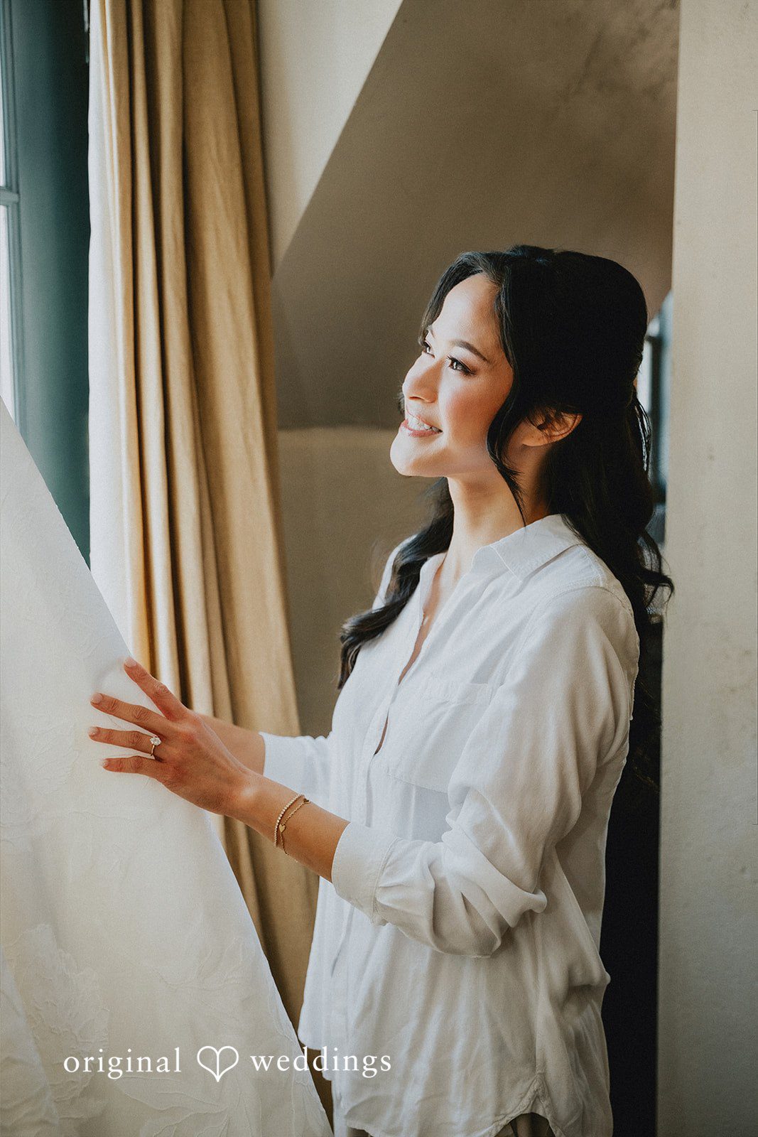 Bride wearing a white silk robe looking out a window.