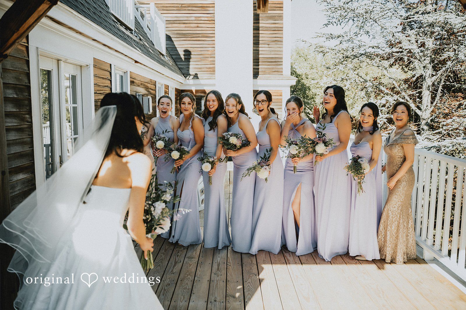 Bride showing her wedding dress to bridesmaids in lavender gowns during a celebratory reveal on a wooden deck.
