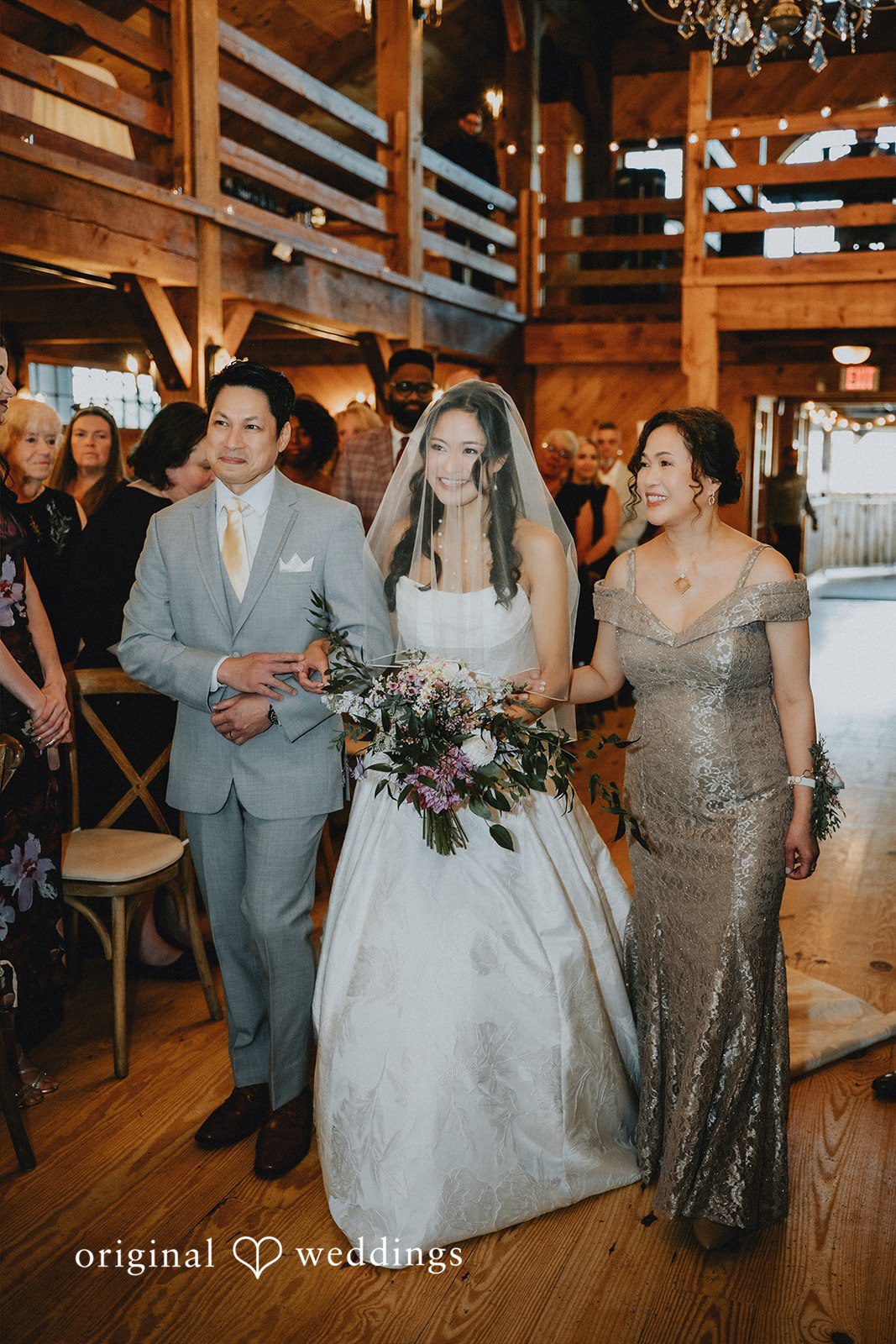 Bride walking down the aisle flanked by two individuals inside a rustic wooden barn venue.