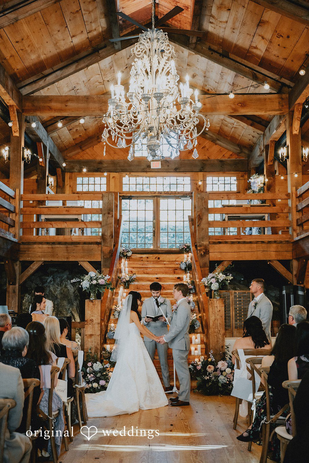 •  Bride and groom holding hands at the altar during a ceremony under a large crystal chandelier inside a wooden barn.