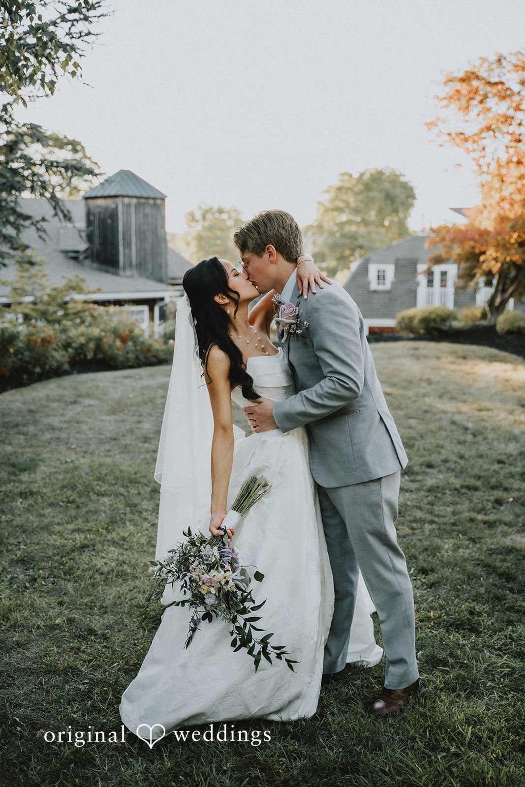 Bride and groom walking and kissing side-by-side across a grassy field.