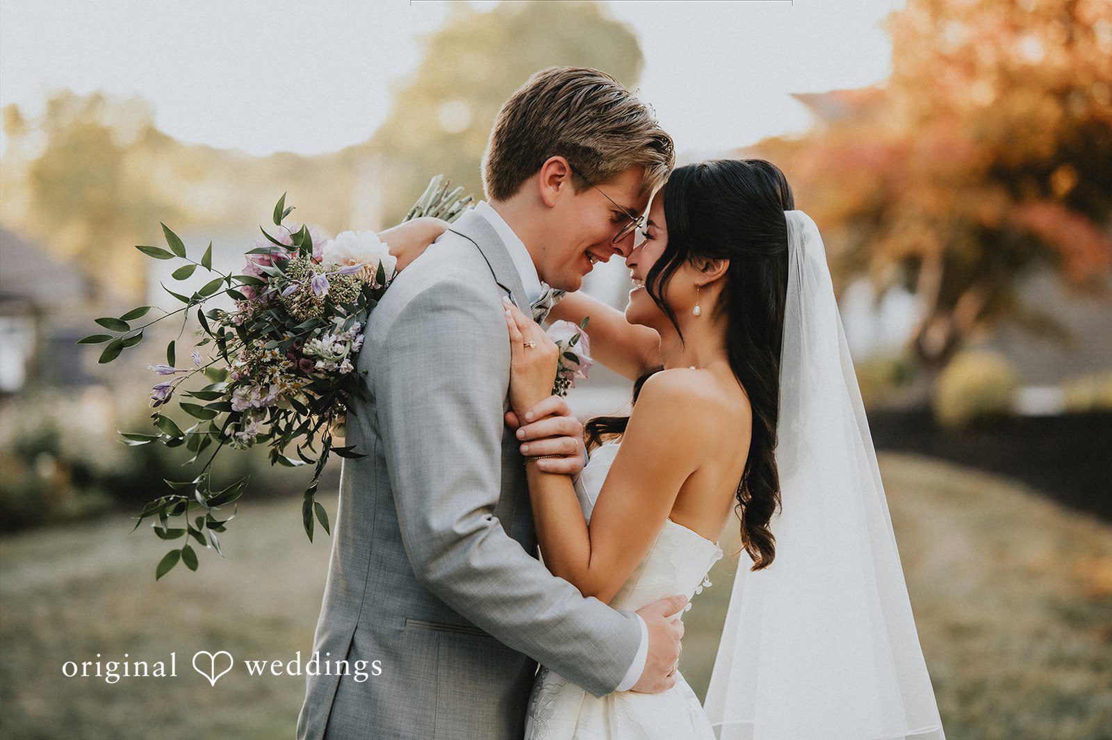 Newlyweds embracing closely and smiling while holding a bouquet of wildflowers on a grassy lawn.