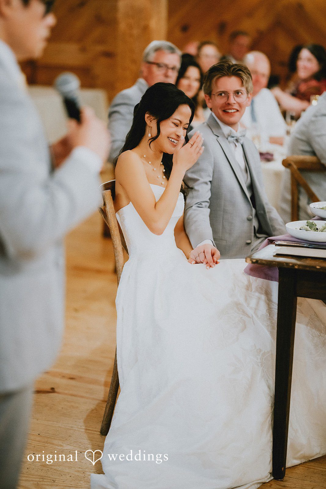Bride sitting at a reception table smiling as wedding guests applaud.