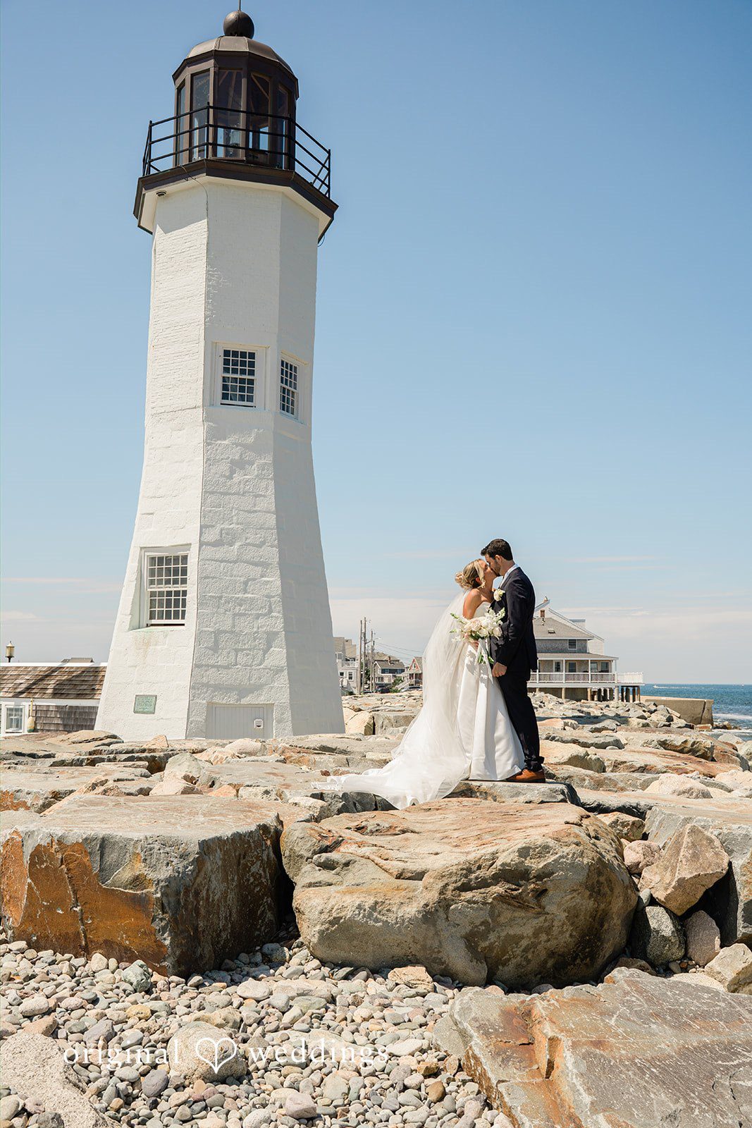 Jessica + Keegan A distant view of a bride and groom standing on a rocky shoreline next to a lighthouse.