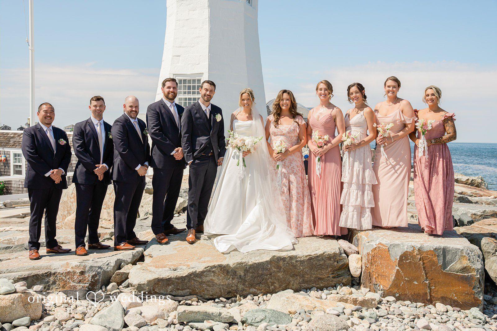 Jessica + Keegan A full wedding party standing in a line on a rocky coastline for a group photo.