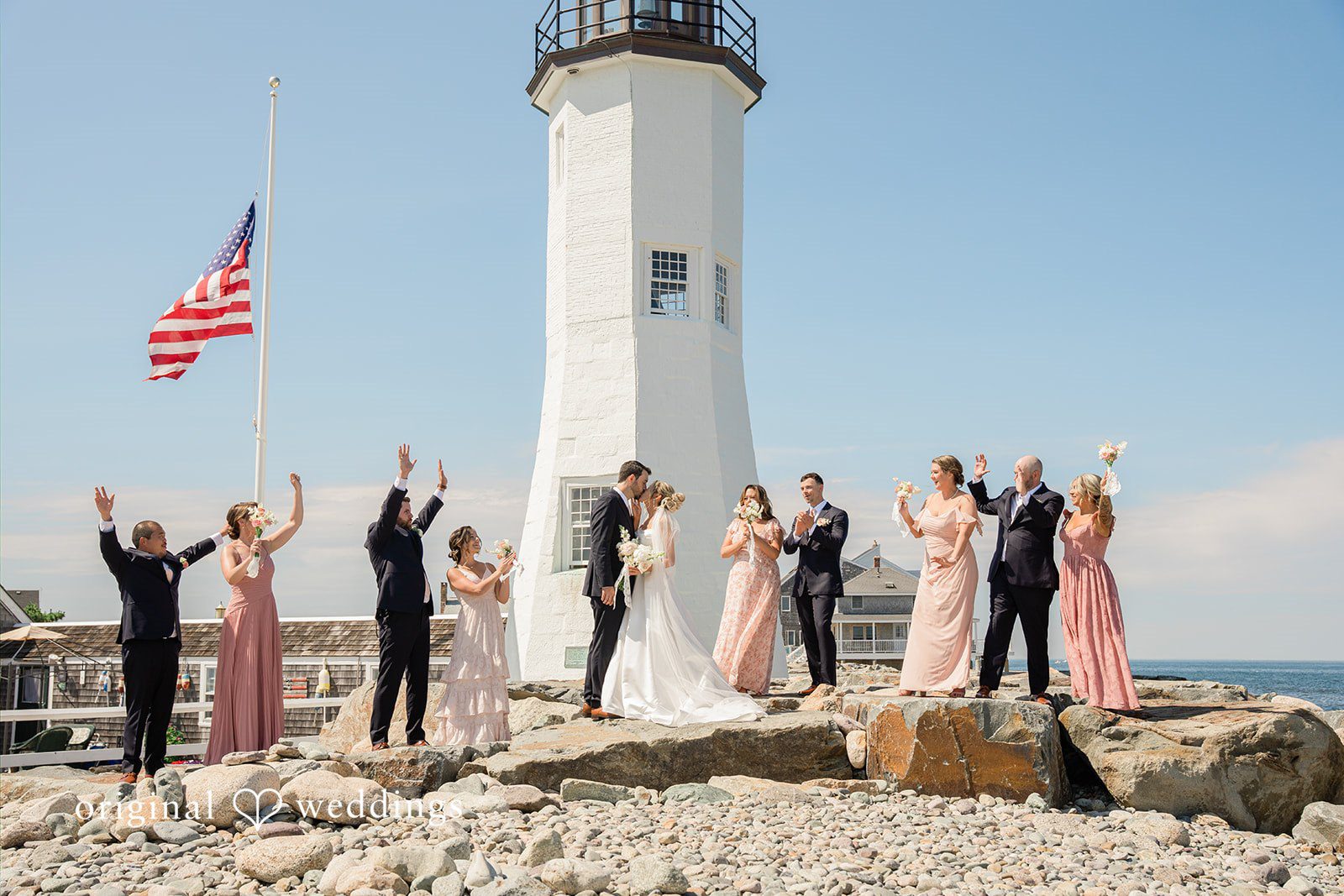 Jessica + Keegan A wedding party posing together on a rocky shore beneath a tall white lighthouse.