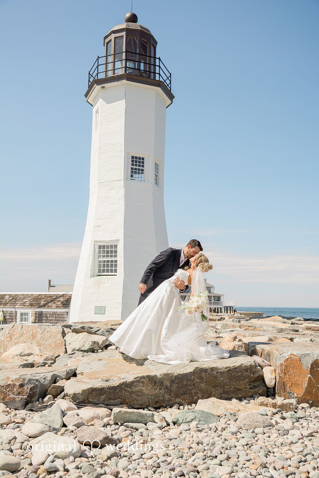 Jessica + Keegan A bride and groom sharing a dip and kiss on a rocky shoreline next to a white lighthouse.