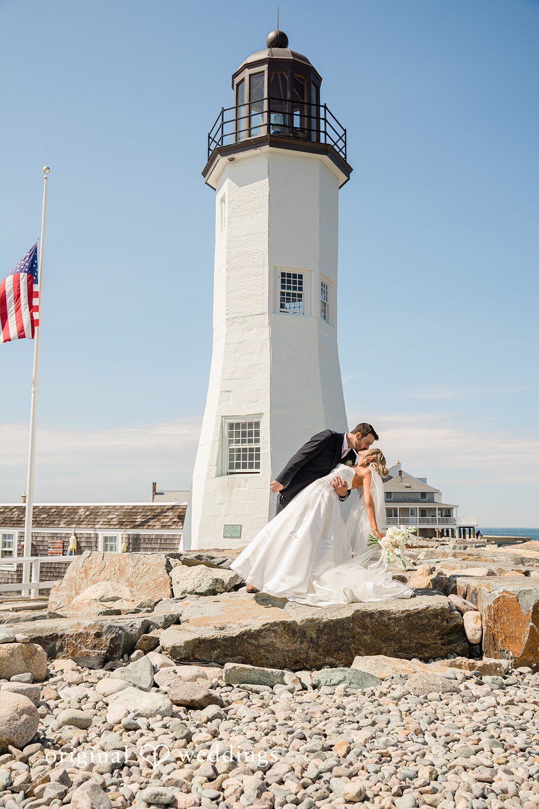 Jessica + Keegan A bride and groom walking together on a rocky beach with a lighthouse in the background.
