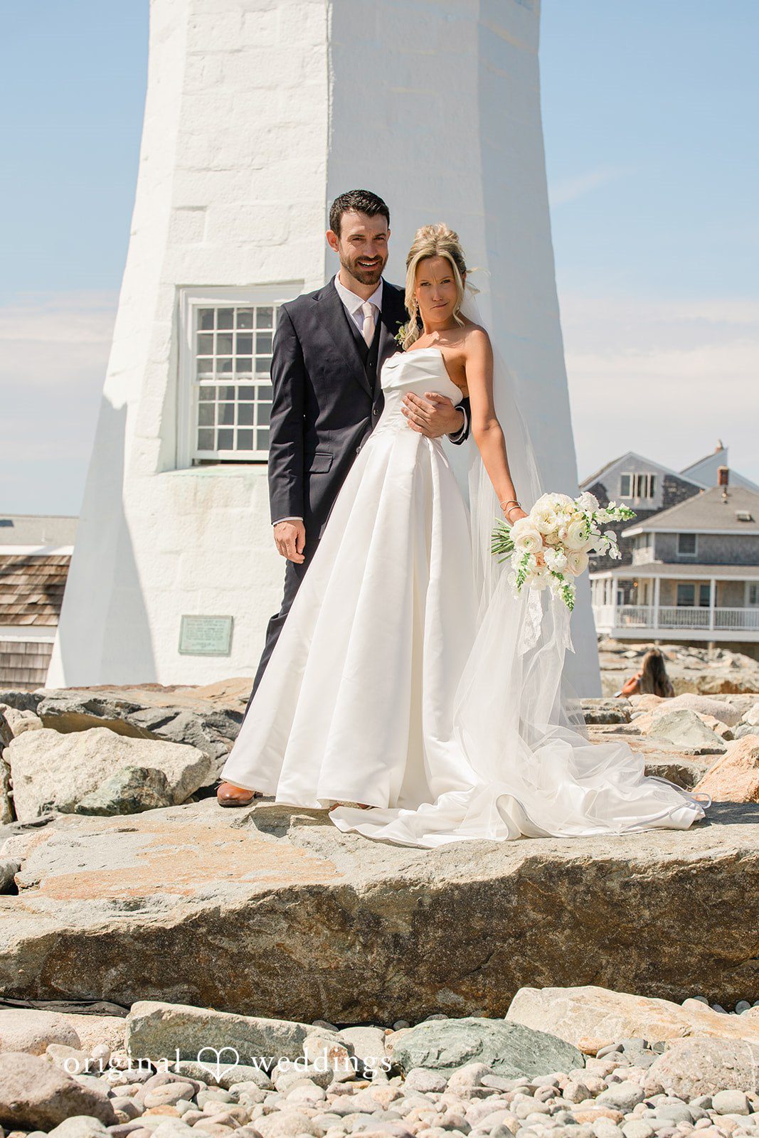 Jessica + Keegan A groom looking at his bride as they stand together near a coastal lighthouse.