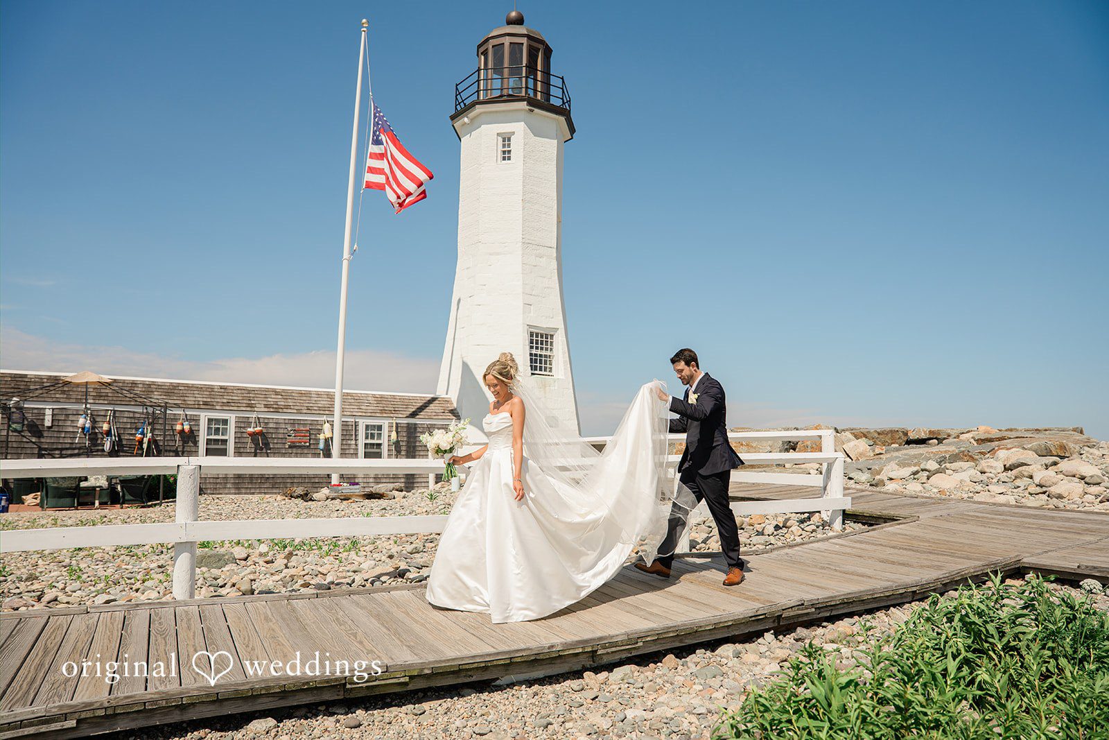 Jessica + Keegan A bride and groom posing on a path with a tall lighthouse stretching into the blue sky.