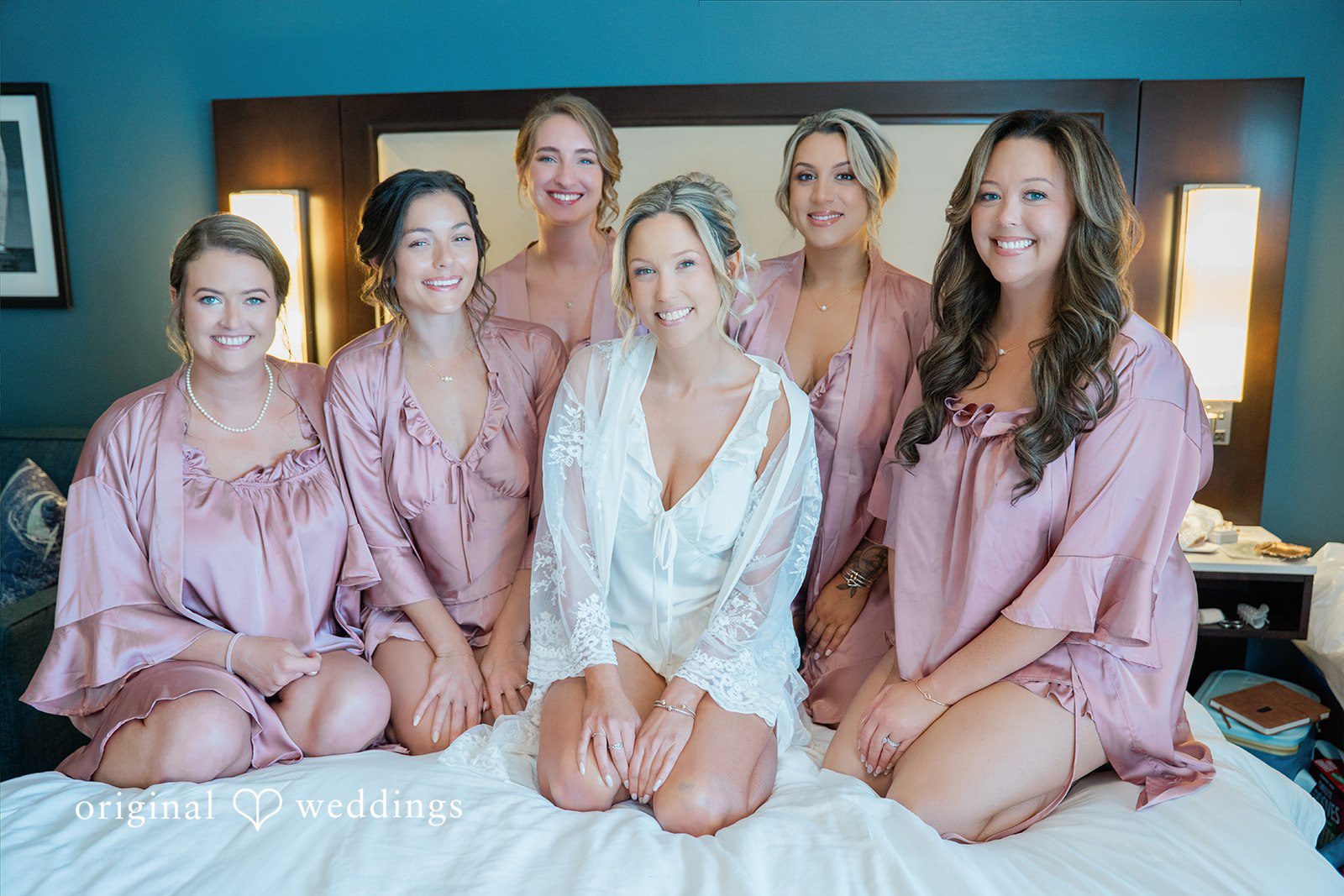 Jessica + Keegan A bride and her bridesmaids in matching silk robes sitting together on a bed.