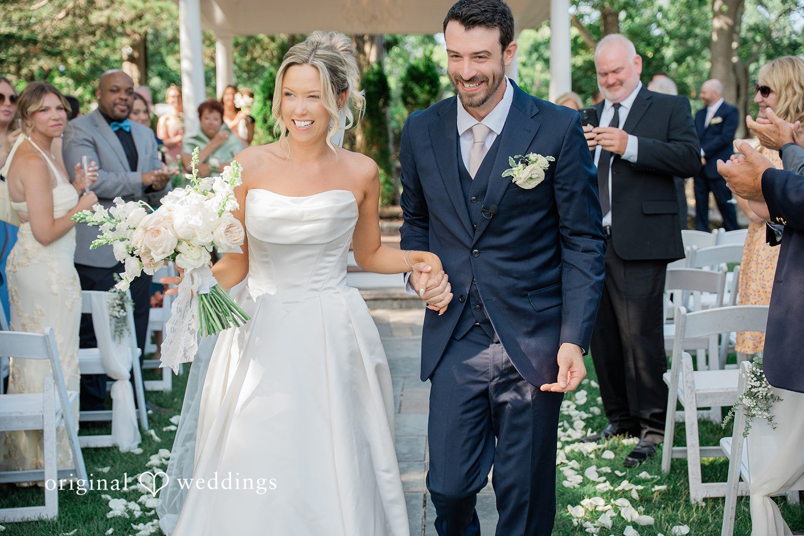 Jessica + Keegan A smiling bride and groom walking down the aisle together as guests look on.