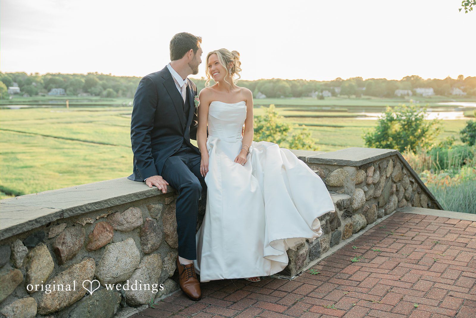 Jessica + Keegan A bride and groom posing together on a stone wall overlooking a scenic landscape.