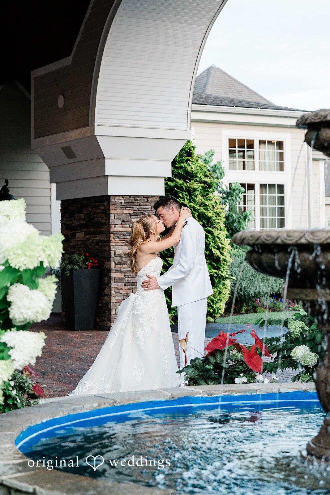 Veronica + Wyatt A bride in a white gown and a groom in a white uniform embracing by a stone fountain.