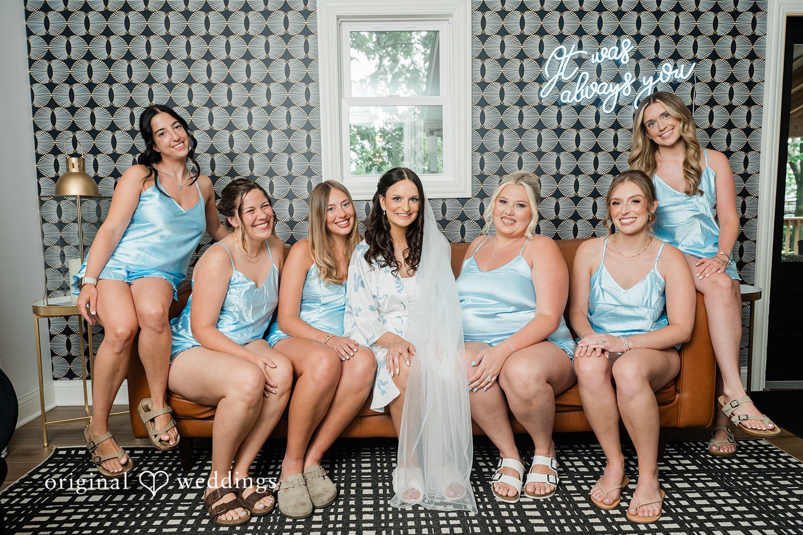 Paige + Stephen Bride and bridesmaids in short light blue dresses sitting together on a dark couch and smiling.