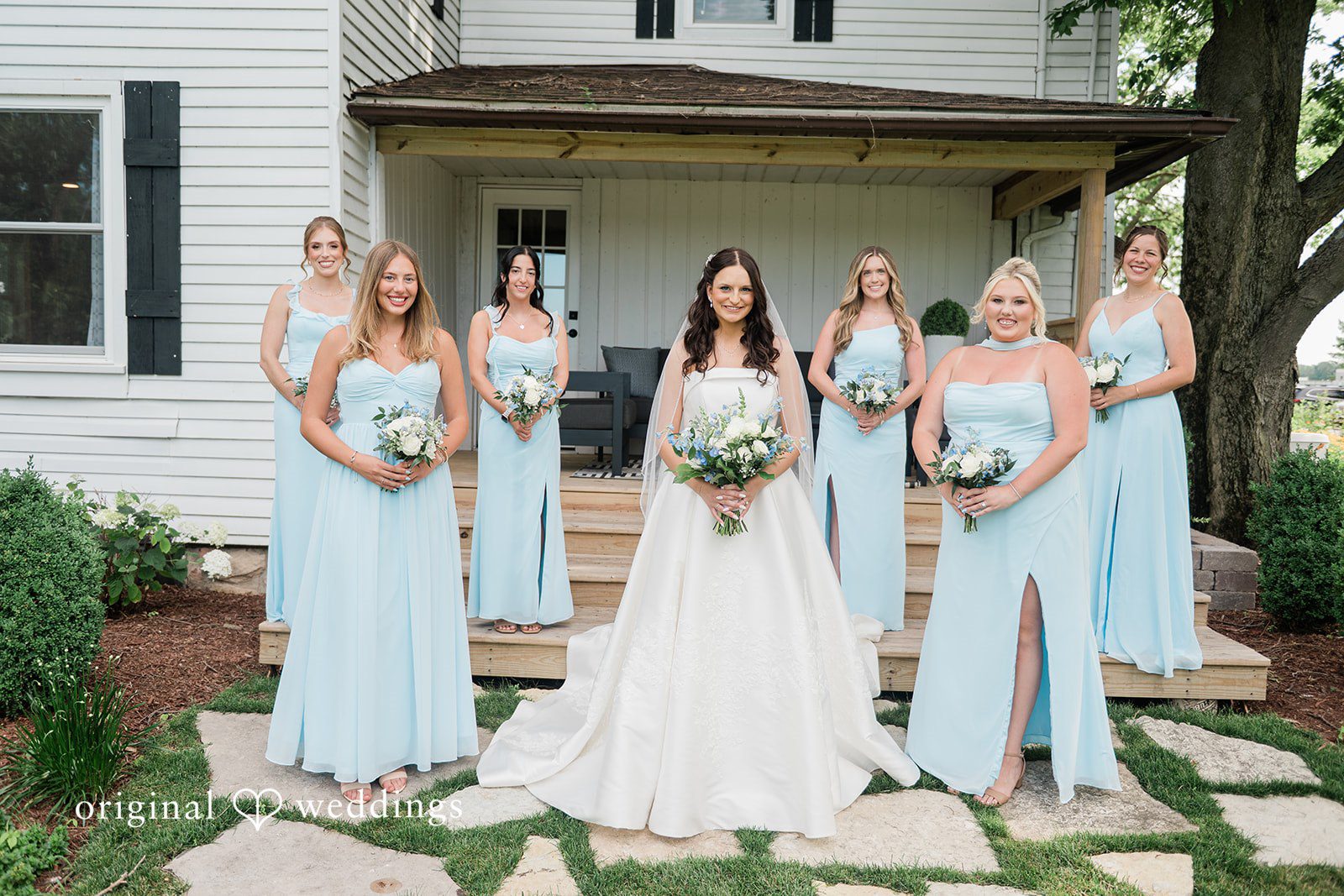 Paige + Stephen A bride and bridesmaids in light blue dresses walking down stone steps outside a white farmhouse.
