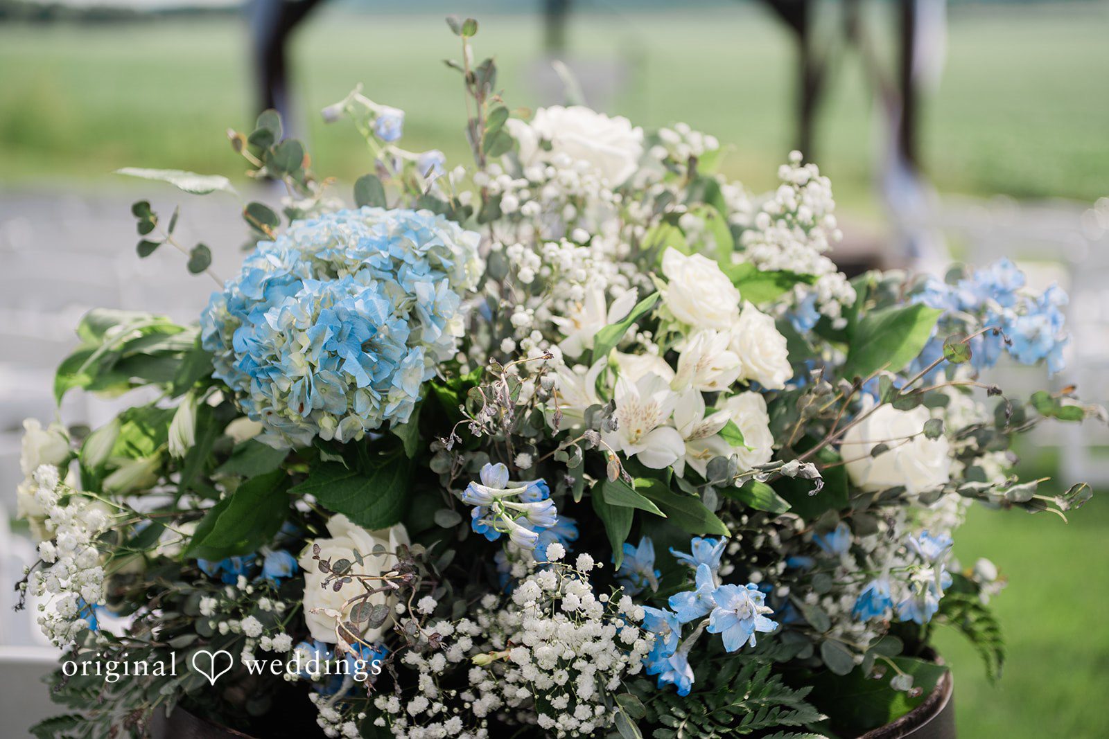 Paige + Stephen A lush floral arrangement featuring white roses, light blue hydrangeas, and trailing green leaves in a rustic vase.