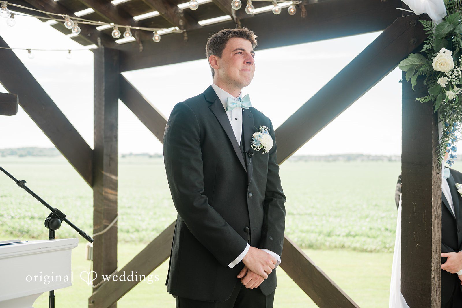 Paige + Stephen A groom in a black suit and bowtie standing next to a dark wooden beam structures.