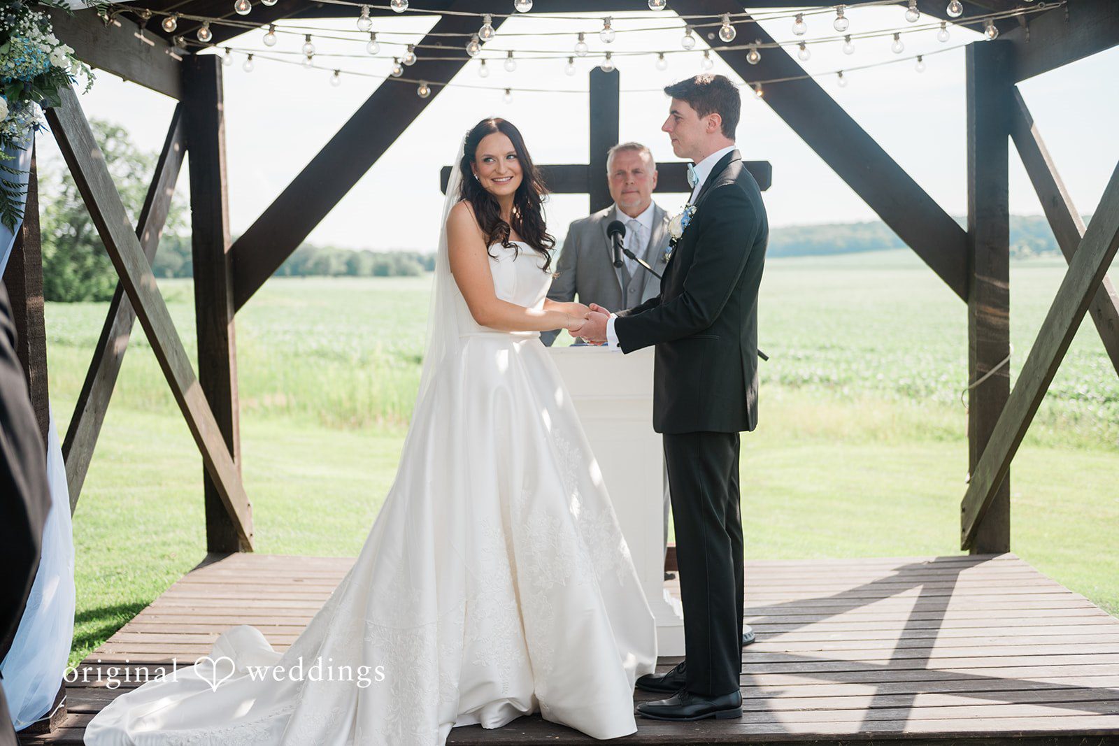 Paige + Stephen A bride and groom standing together beneath a dark wooden pergola structure in a grassy field.