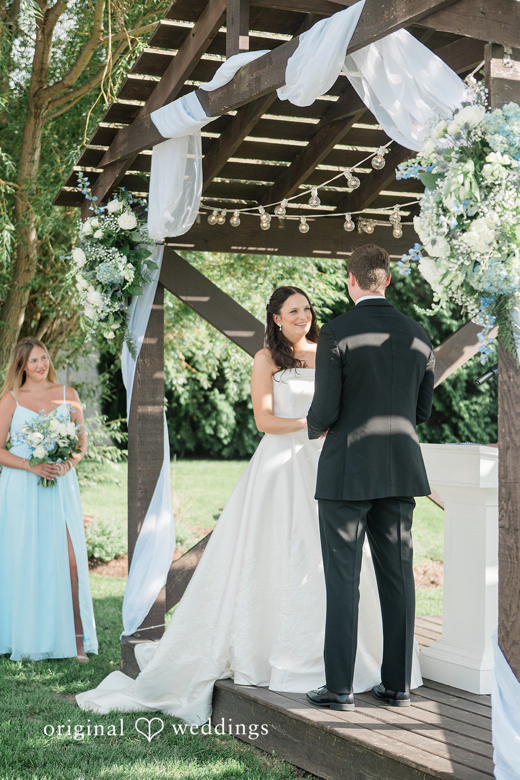 Paige + Stephen A bride in a long gown walking toward a groom standing beneath trees during a ceremony.