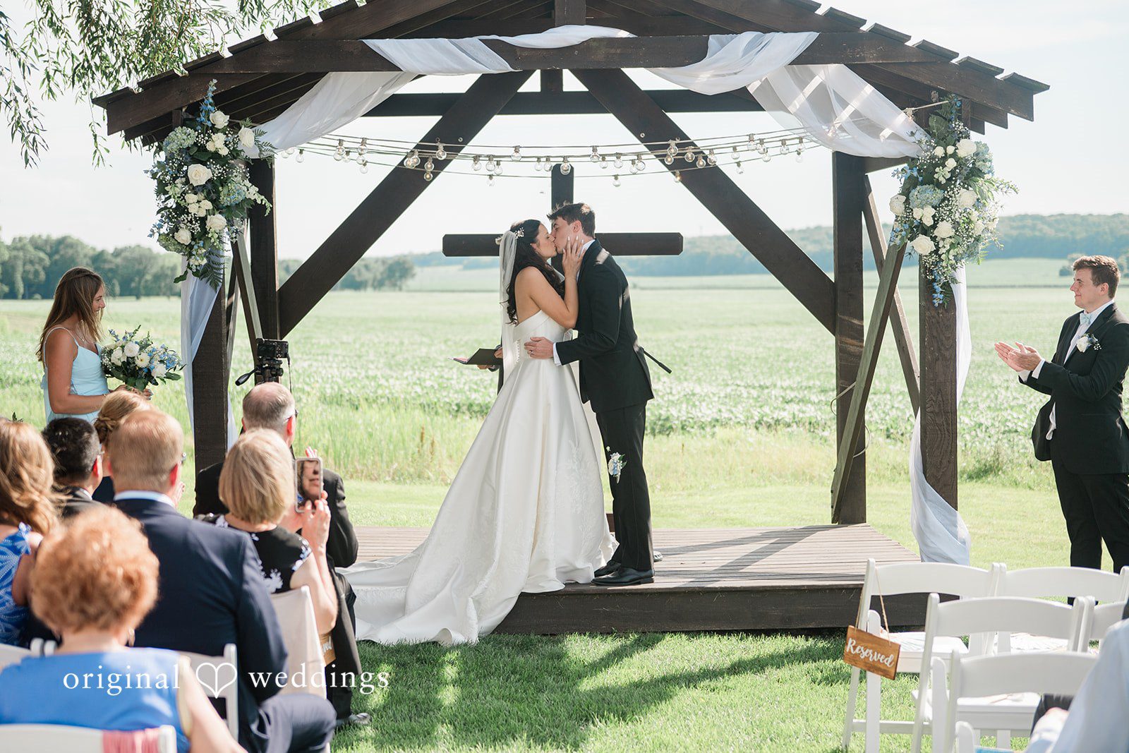 Paige + Stephen A wide shot of a wedding ceremony under a wooden archway with guests looking on from a grassy field.