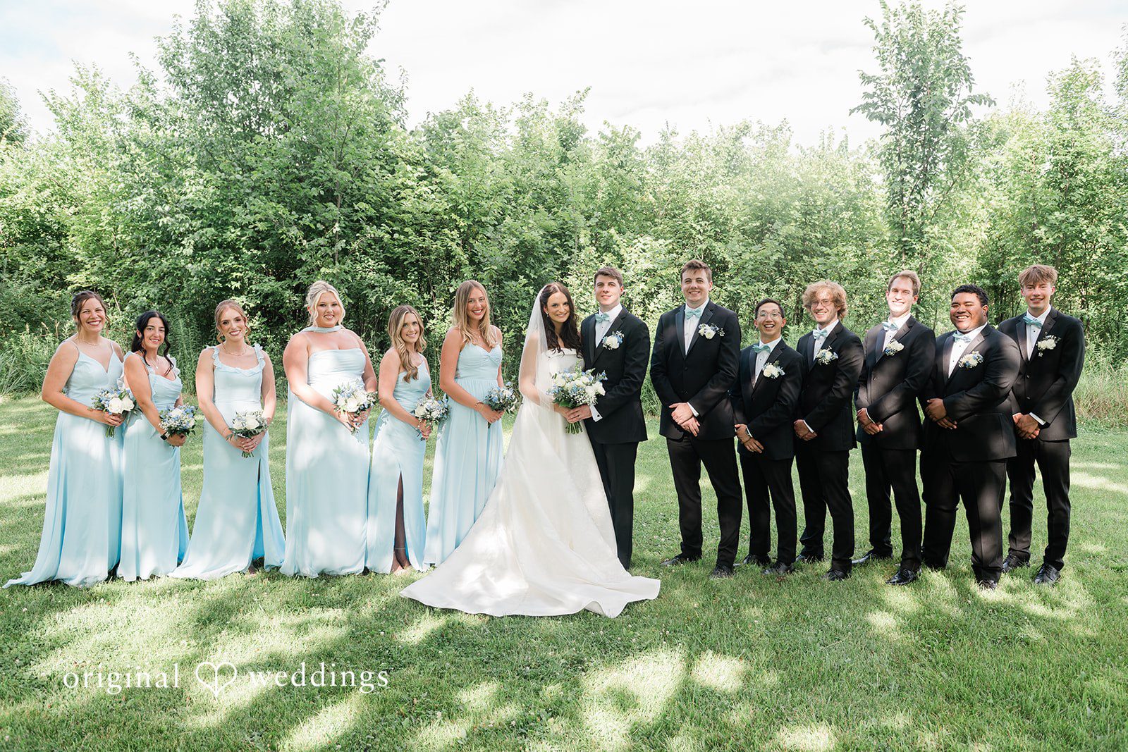 Paige + Stephen A large wedding party with bridesmaids in light blue and groomsmen in black suits posing in a field.