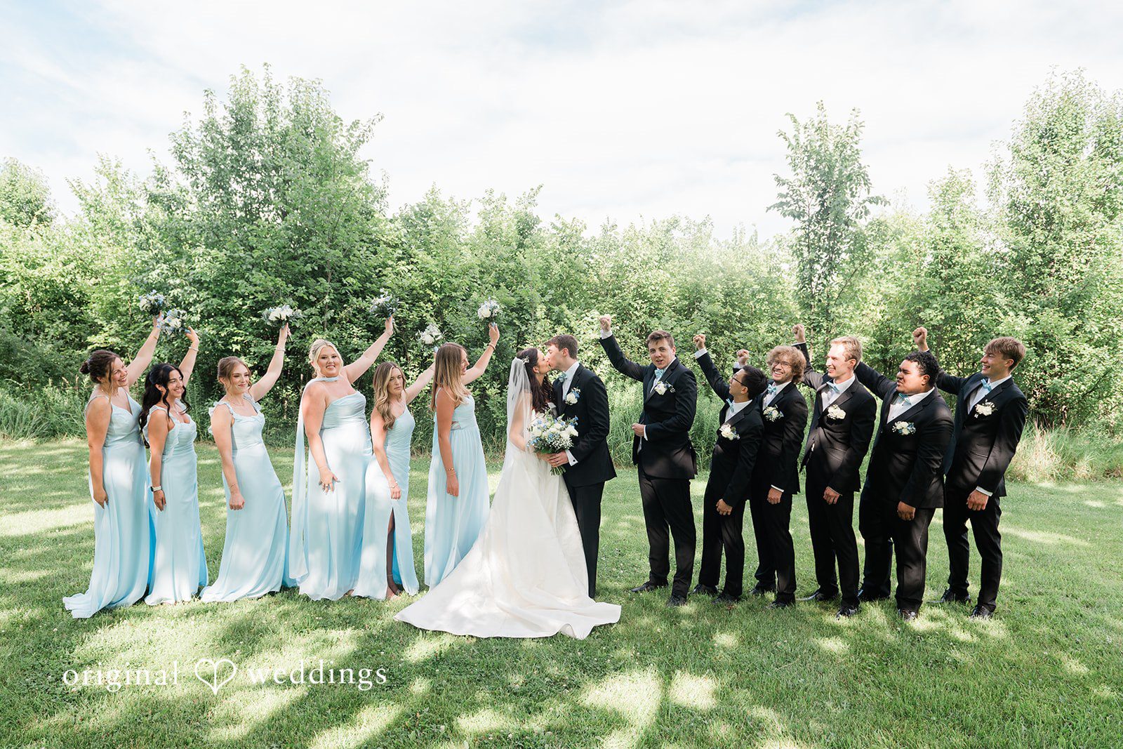 Paige + Stephen A wide shot of a full wedding party standing together for a photo in a green park.