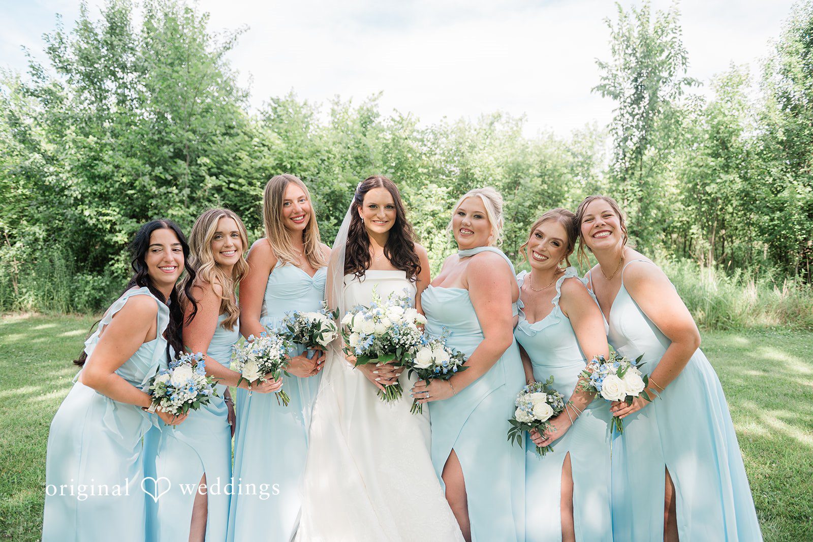 Paige + Stephen Bride and bridesmaids in light blue dresses holding matching bouquets while standing together in a grassy area.