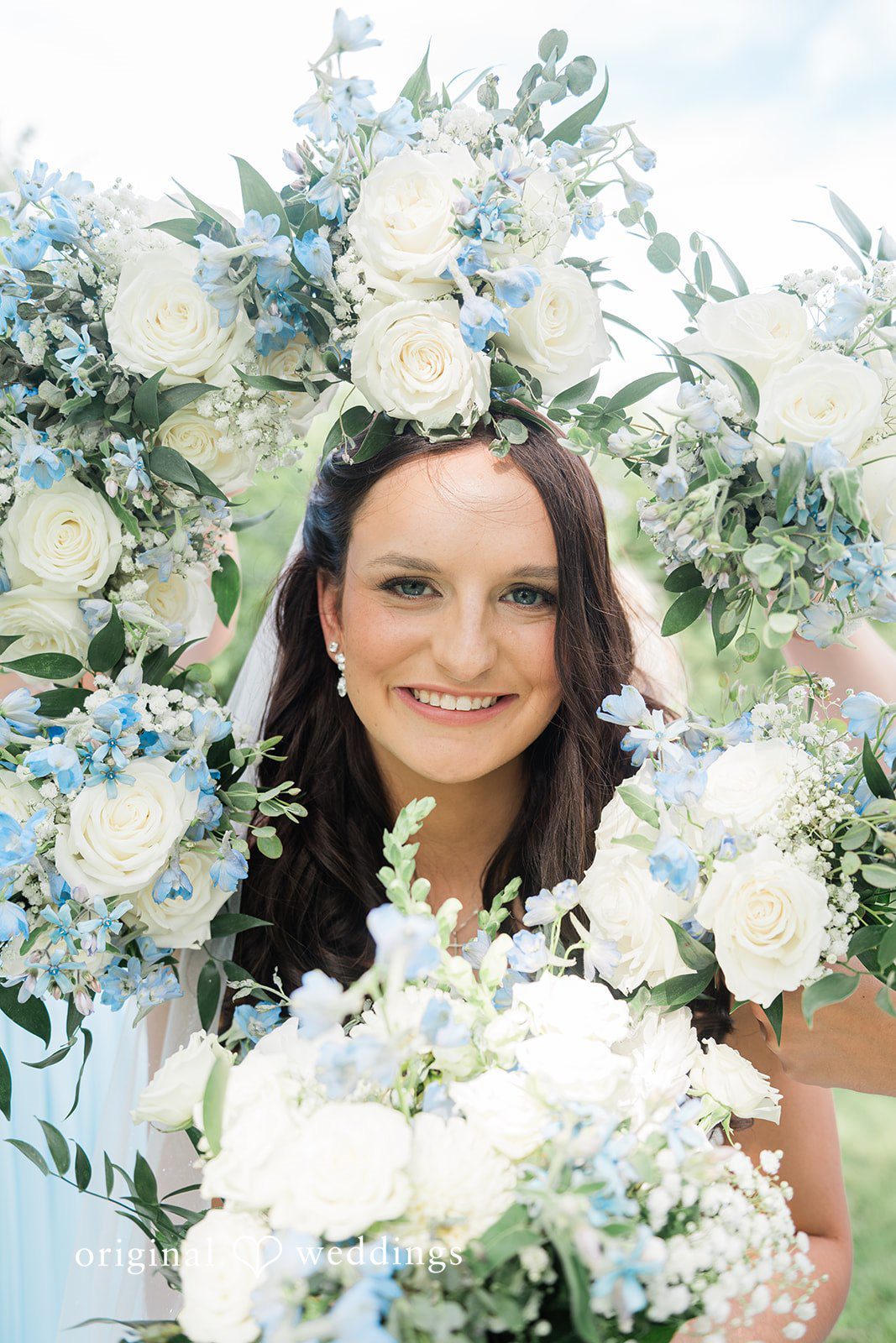Paige + Stephen A close-up portrait of a smiling bride with a floral crown, surrounded by white flowers and green leaves.