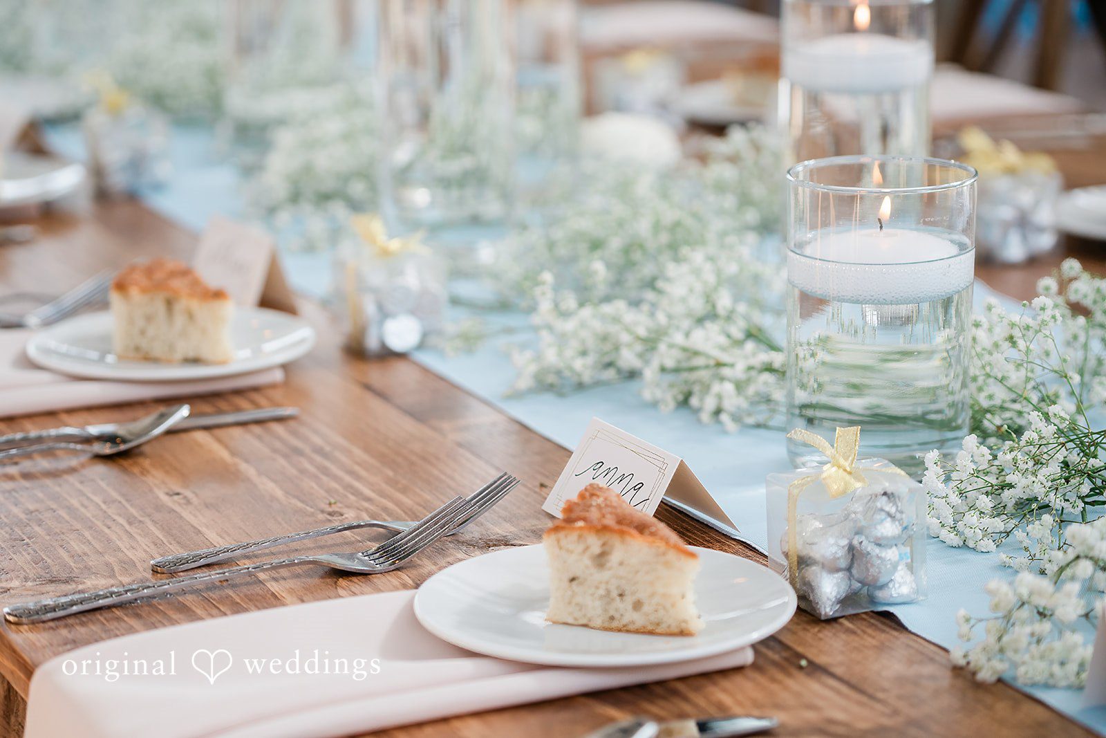 Paige + Stephen A close-up view of a rustic wooden tabletop decorated with delicate white lace, glass candleholders, and light blue florals.